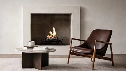 Modern minimalist living room featuring a built-in fireplace with a clean-burning flame set in a recessed concrete surround. In front, a round stone coffee table with books and a white mug sits on a textured rug beside a mid-century style brown leather lounge chair with wooden arms, creating a warm, contemporary interior setting.