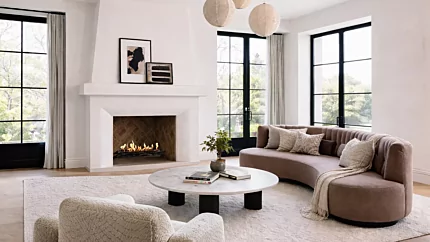 EcoSmart Fire Heritage 50 Grate installed in a white built-in fireplace with visible flame, set in a bright contemporary living room featuring a curved sofa, marble coffee table and large black-framed windows.